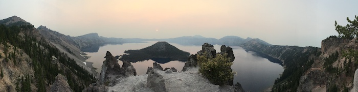 Panoramic of Crater Lake, a scenic view of water, mountains, trees and hazy sun.